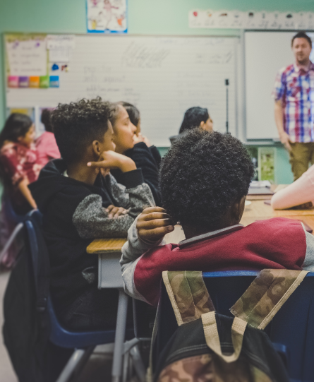 A male teacher stands in front of his classroom of teenage children, many of color.